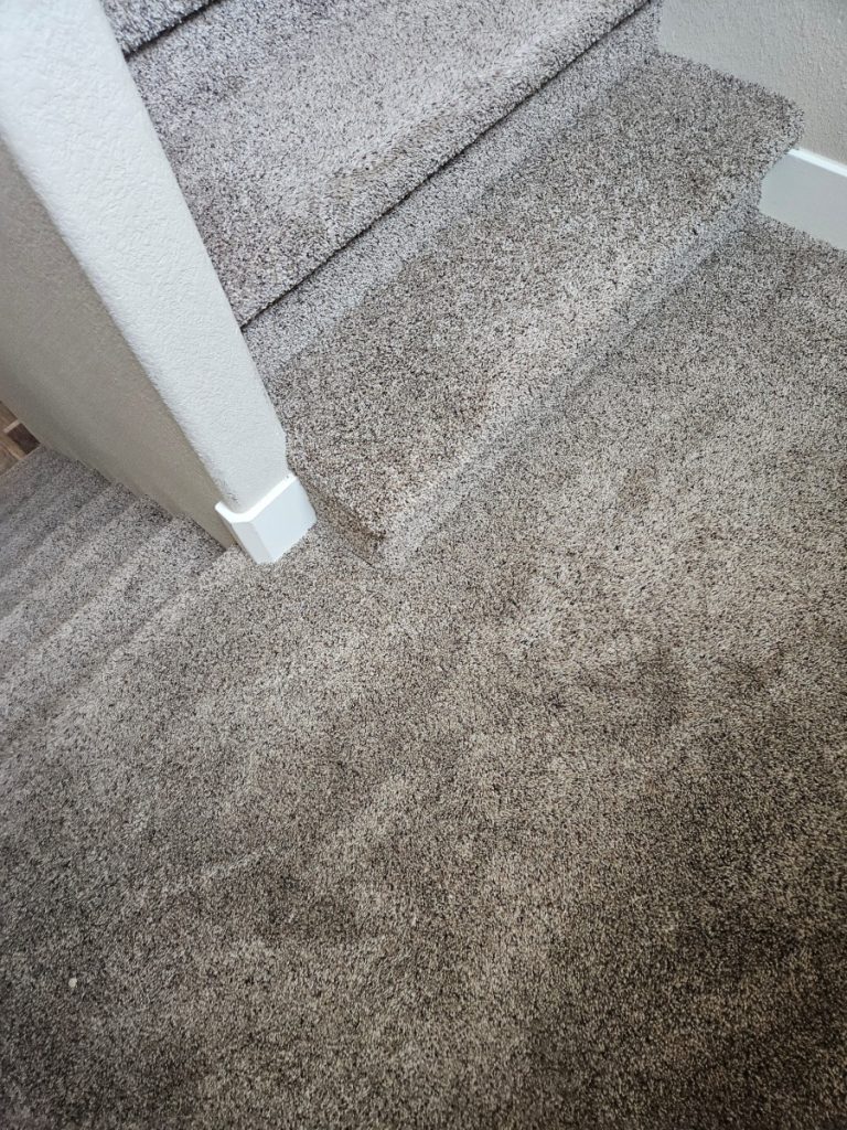 A close-up view of carpeted stairs and floor, featuring light brown textured carpeting, white baseboards, and light-colored walls.