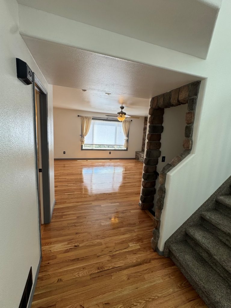 A hallway with shiny hardwood floors leads to a bright living room with large windows, white curtains, a ceiling fan, and a stone-trimmed arch near carpeted stairs.